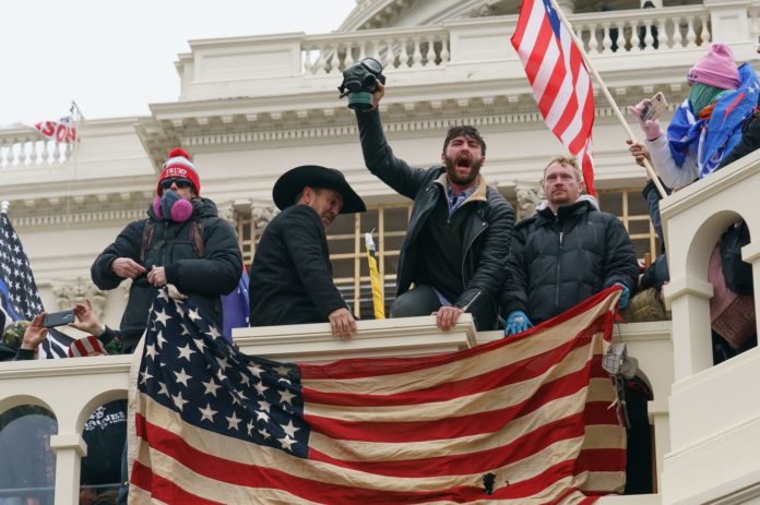 Rioters outside the capitol building on Jan 6 holding american flags and sporting gas masks, seemingly yelling and chanting together.