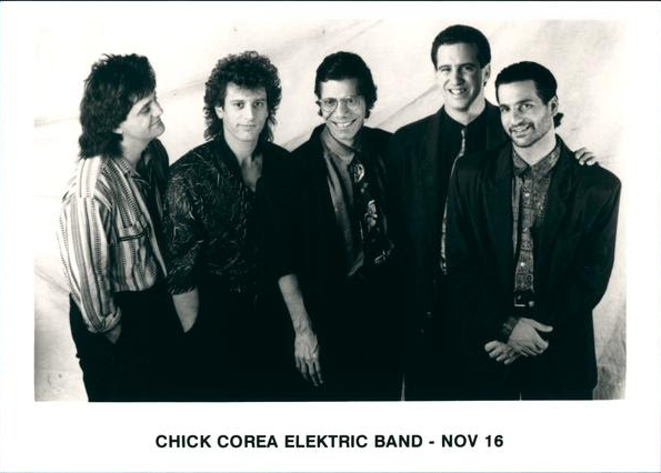 Members of the Chick Corea Jazz Electric Band standing next to one another smiling with a white background, all wearing dark clothing