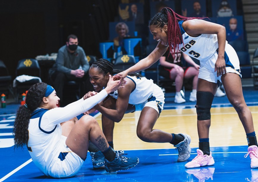 Three UMKC womens basketball players, one on the ground and two standing helping the third one off the ground all smiling and cheerful