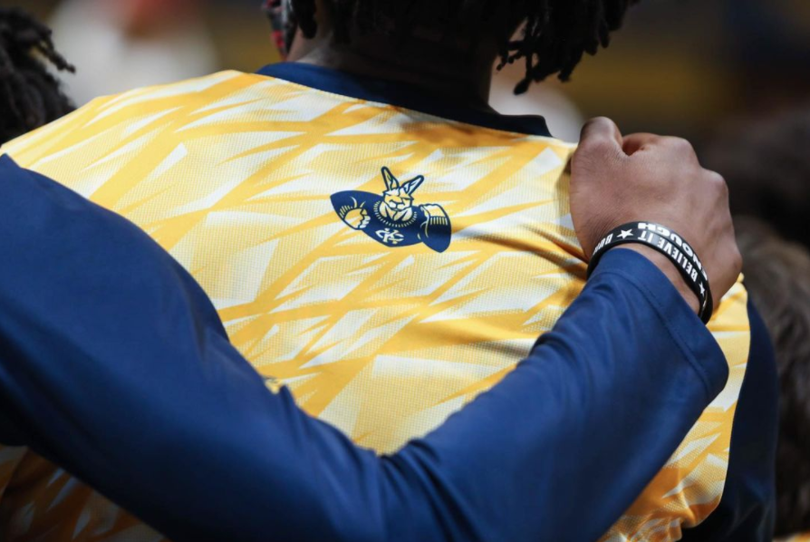 Back of a UMKC Mens Basketball practice jersey with yellow and the UMKC kangaroo on the back. There is an arm wrapped around the shoulders of another player