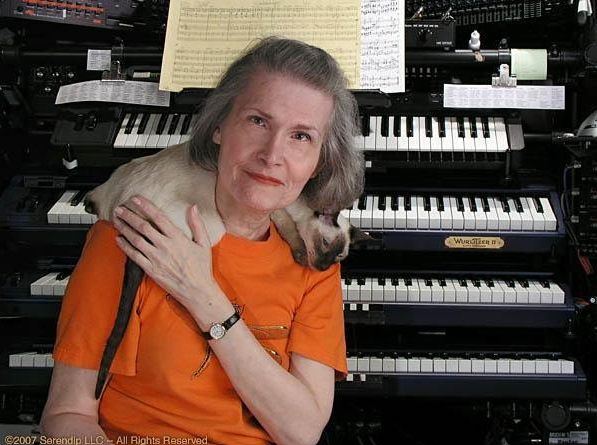 Pianist Wendy Carlos sitting in front of her piano with her Siamese cat sleeping on her shoulders